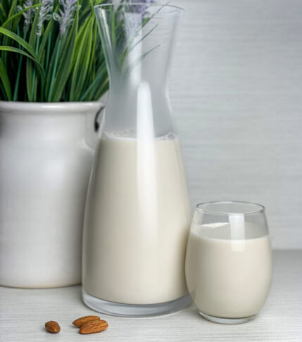 Homemade almond milk in a glass cup next to a carafe on a rustic kitchen table.