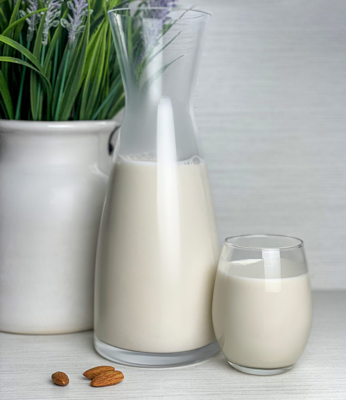 Homemade almond milk in a glass cup next to a carafe on a rustic kitchen table.