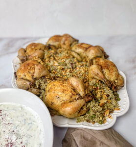 A beautifully plated dish of stuffed Cornish hens with rice and lamb, garnished with peas and served alongside a refreshing yogurt cucumber salad.