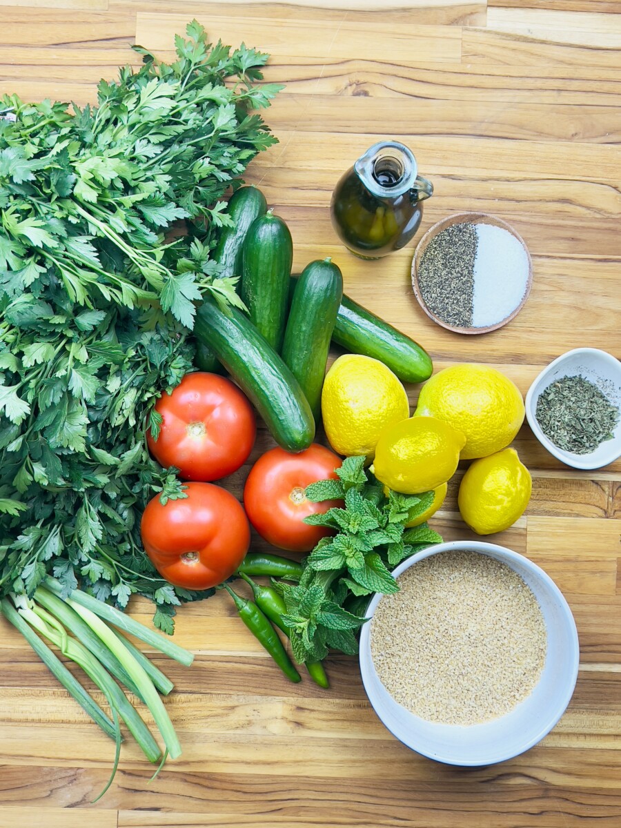 Fresh ingredients for tabbouleh laid out on a board: parsley, mint, bulgur, tomatoes, green onions, lemon, and olive oil.
