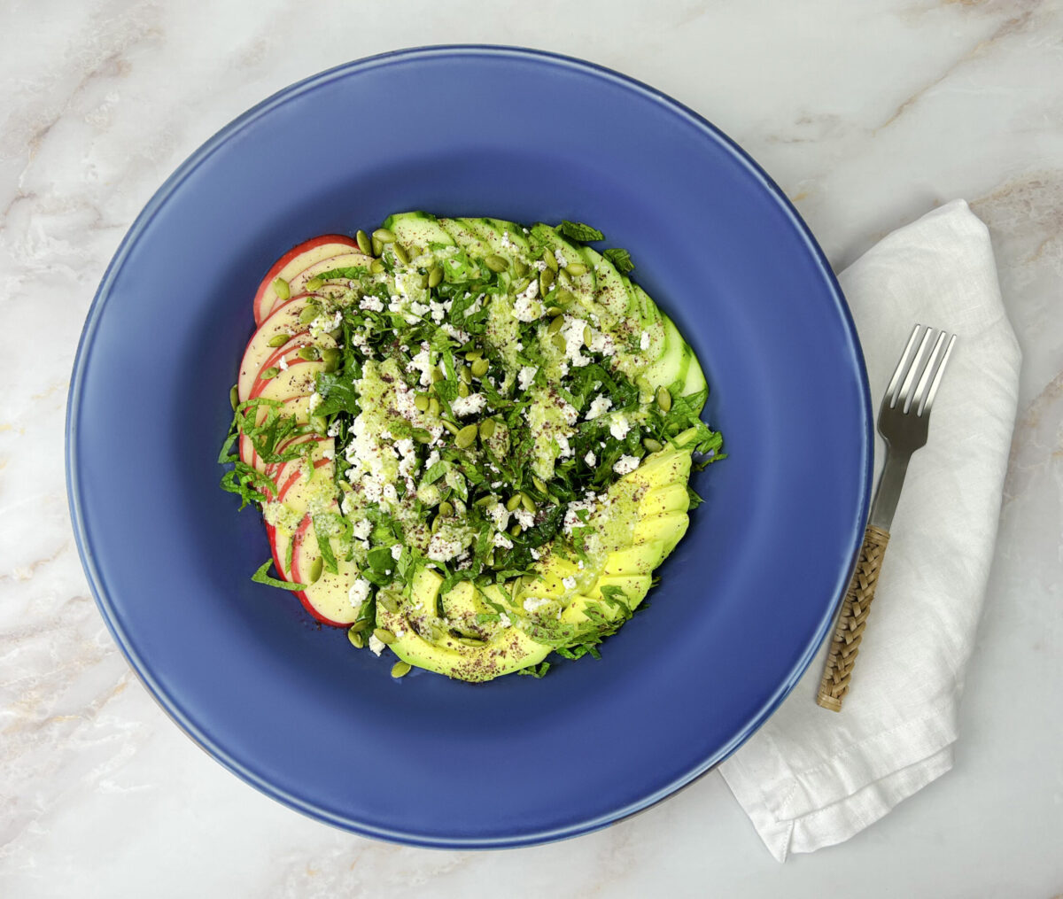 A vibrant and nutritious kale apple salad with slices of cucumber, avocado, apple, sprinkled with feta cheese, toasted pumpkin seeds, and a hint of sumac."