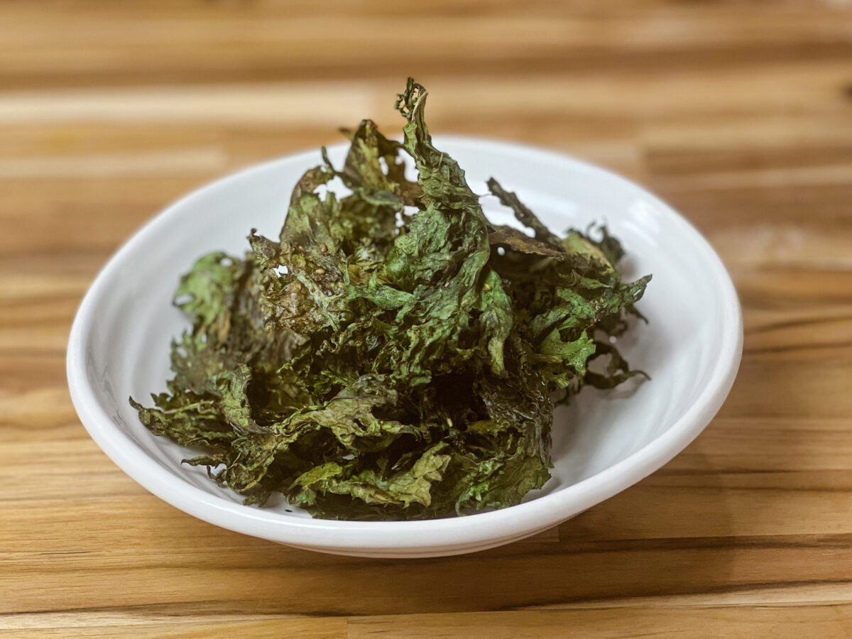 A bowl of crispy za'atar-seasoned kale chips on a wooden table, garnished with a slice of lemon.