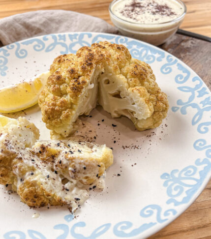 Golden-brown whole roasted cauliflower served with creamy tahini feta sauce, garnished with sumac and nigella seeds.