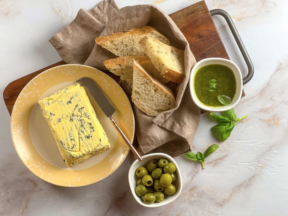A block of fresh basil butter next to some bread and a bowl of olives