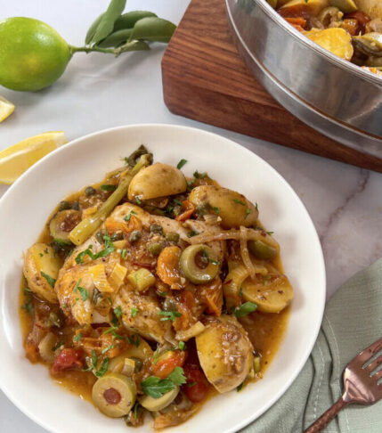 Steaming Chilean Sea Bass on a bed of tomatoes, olives, and potatoes, garnished with fresh herbs.