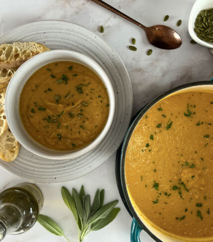 Large pot and bowl of roasted Butter Nut Squash Soup garnished with toasted pumpkin seeds and accompanied by slices of baguette bread