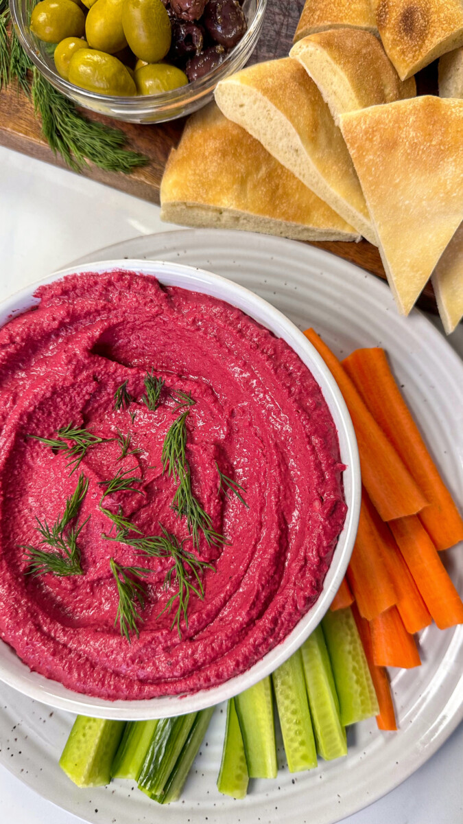 A vibrant bowl of roasted beet hummus garnished with fresh parsley and sprinkled with sesame seeds, served with warm pita bread on a rustic wooden table.