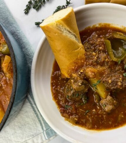 A delicious bowl of homemade beef stew with chunks of tender beef, potatoes, and green peppers, served with a slice of crusty bread soaked in flavorful broth.