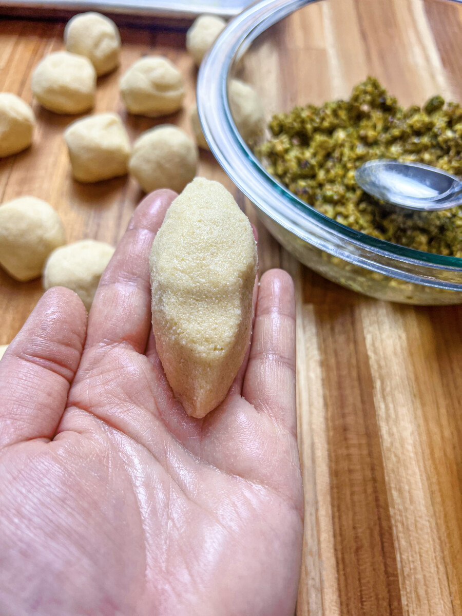 A photo showing a hand shaping a ball of filling into a flattened round or oval shape, surrounded by a sandy mixture of semolina, sugar, clarified butter, and yogurt. The surface of the cookie is being scored with a fork or decorative tool, creating intricate patterns. This traditional method of handcrafting Mammoul cookies is a testament to the skill and patience of Middle Eastern bakers and results in a delicious and visually stunning final product
