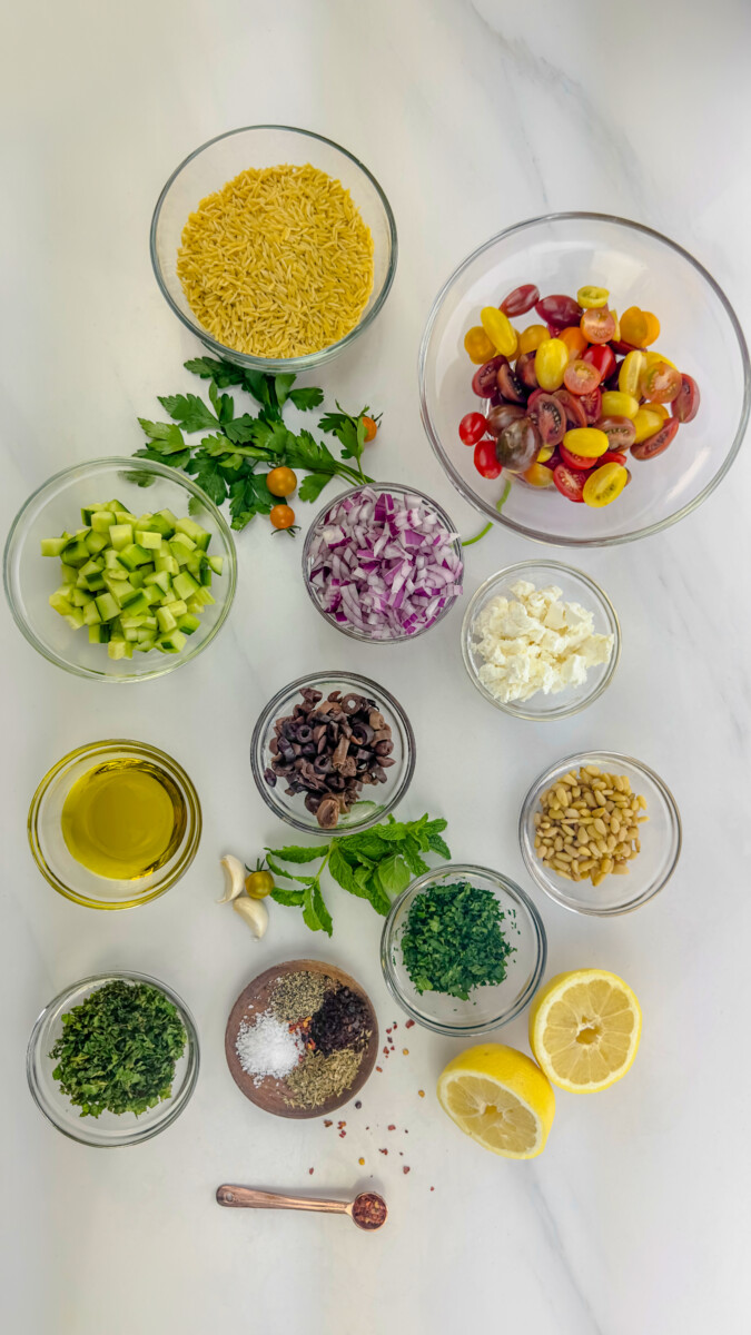 Measured ingredients for Greek Orzo Salad including orzo pasta, cherry tomatoes, cucumber, red onion, Kalamata olives, and fresh herbs.