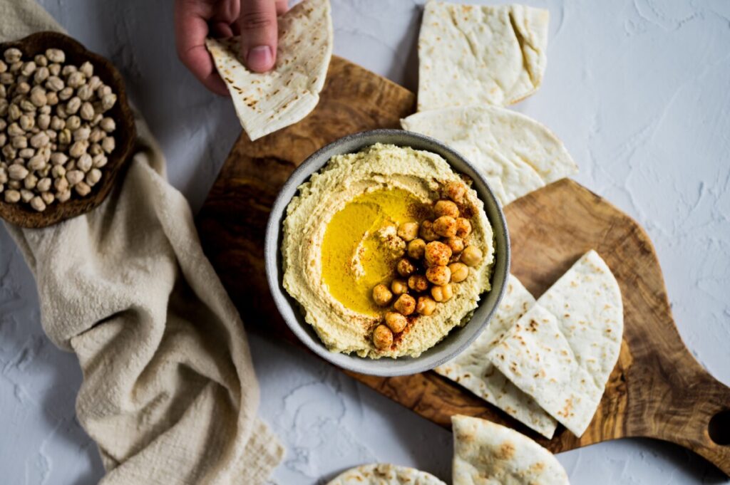 A serving board featuring hummus, pieces of pita bread, and a small bowl of dry chickpeas.