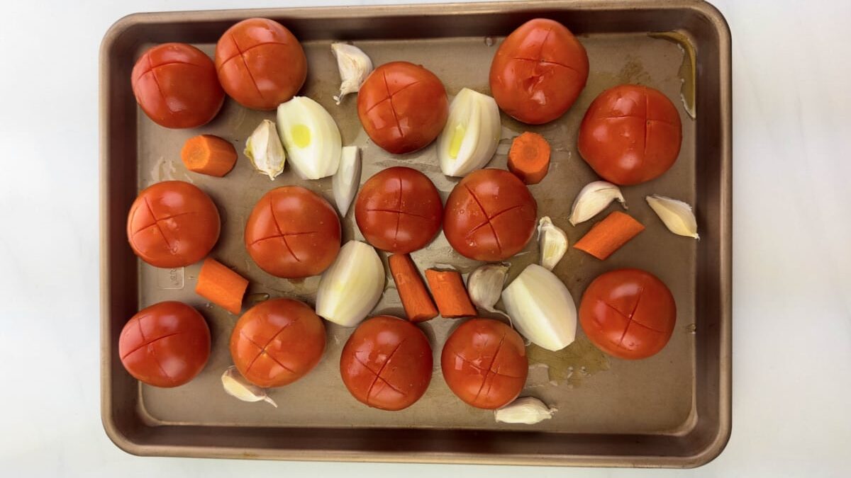 Fresh vegetables on a baking sheet, ready for roasting: tomatoes, onions, garlic, and carrots