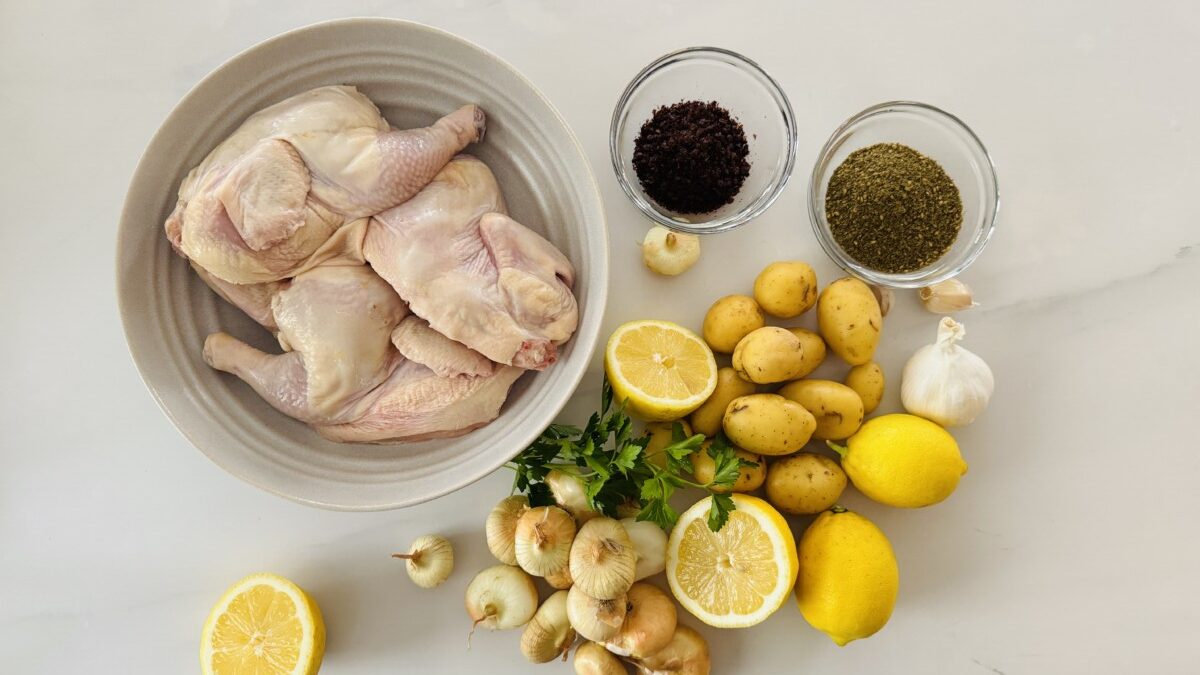 A visual feast of ingredients laid out on a countertop. It includes two halved Cornish hens, a bowl of golden marinade, a bunch of fresh lemons, a container of zaatar spice, a bag of golden potatoes, peeled cipollini onions, and a head of garlic.