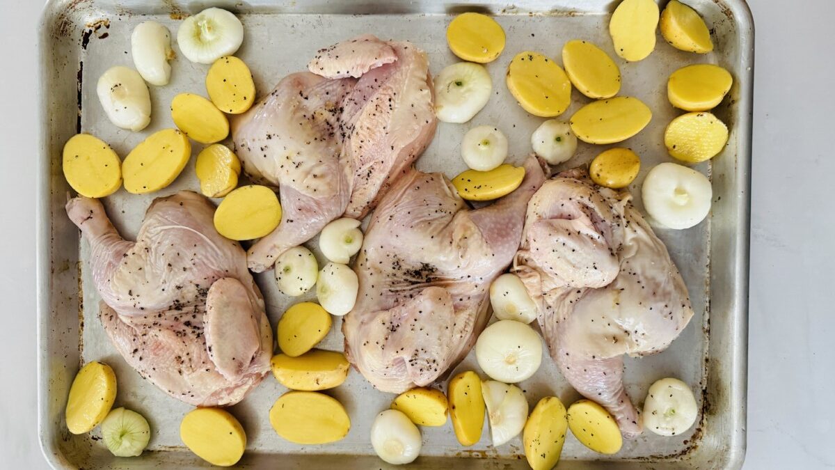 A large baking tray filled with two seasoned Cornish hen halves, surrounded by golden potato wedges and peeled cipollini onions.