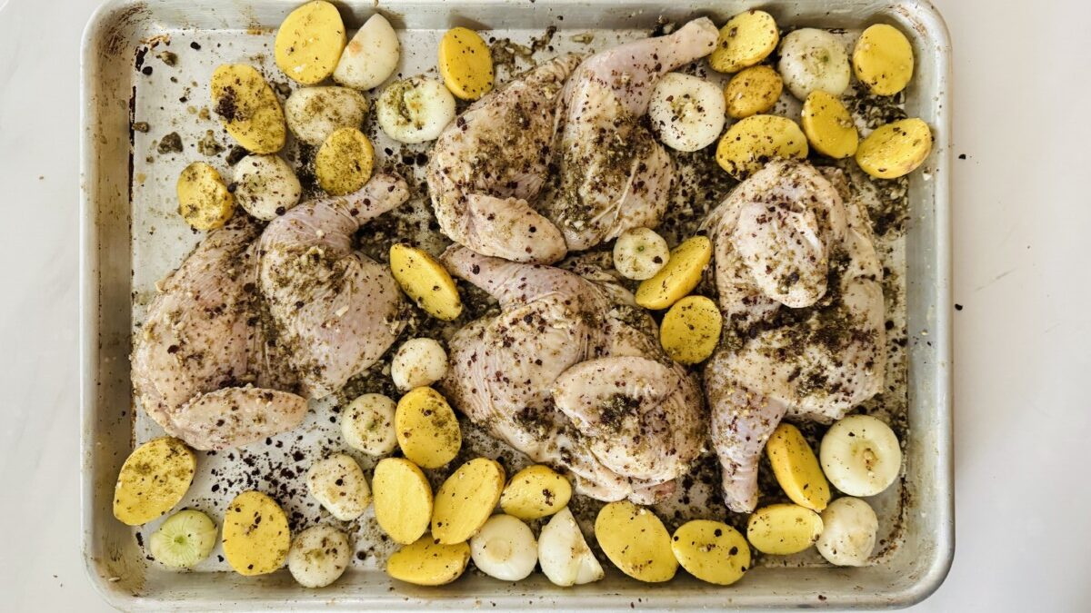 A large baking tray filled with seasoned Cornish hen halves, golden potato wedges, and peeled cipollini onions, bathed in a golden yellow marinade.