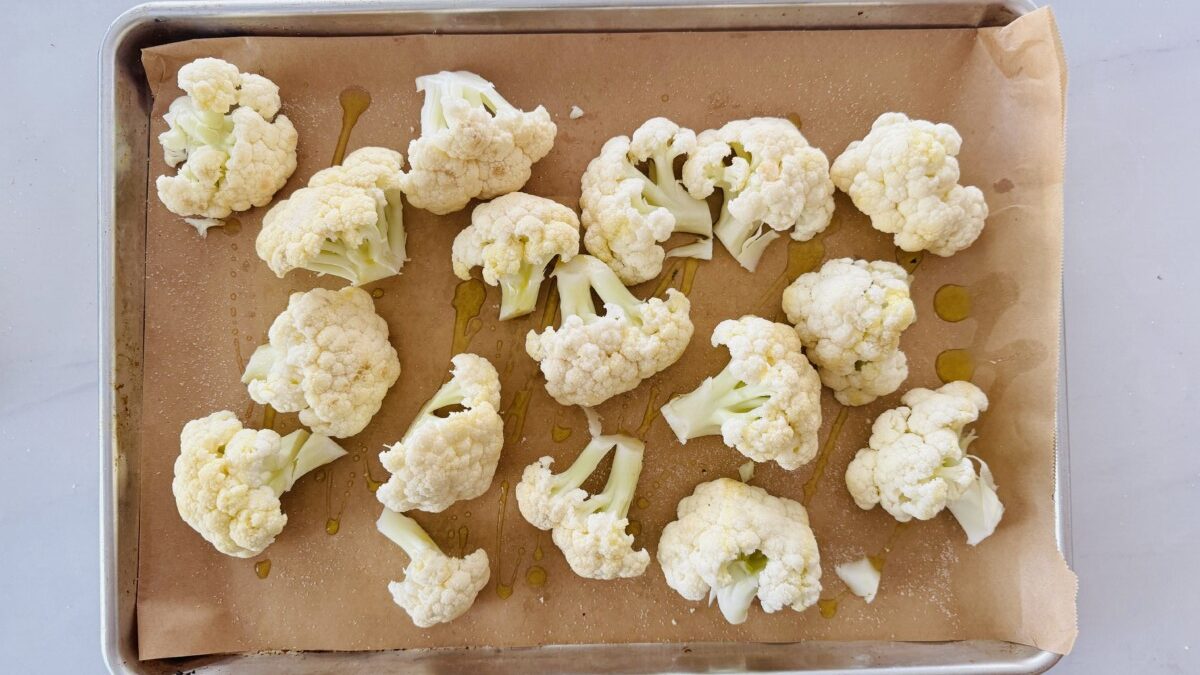 Raw cauliflower florets arranged on a parchment paper-lined baking sheet. A spoon drizzles olive oil over the cauliflower.