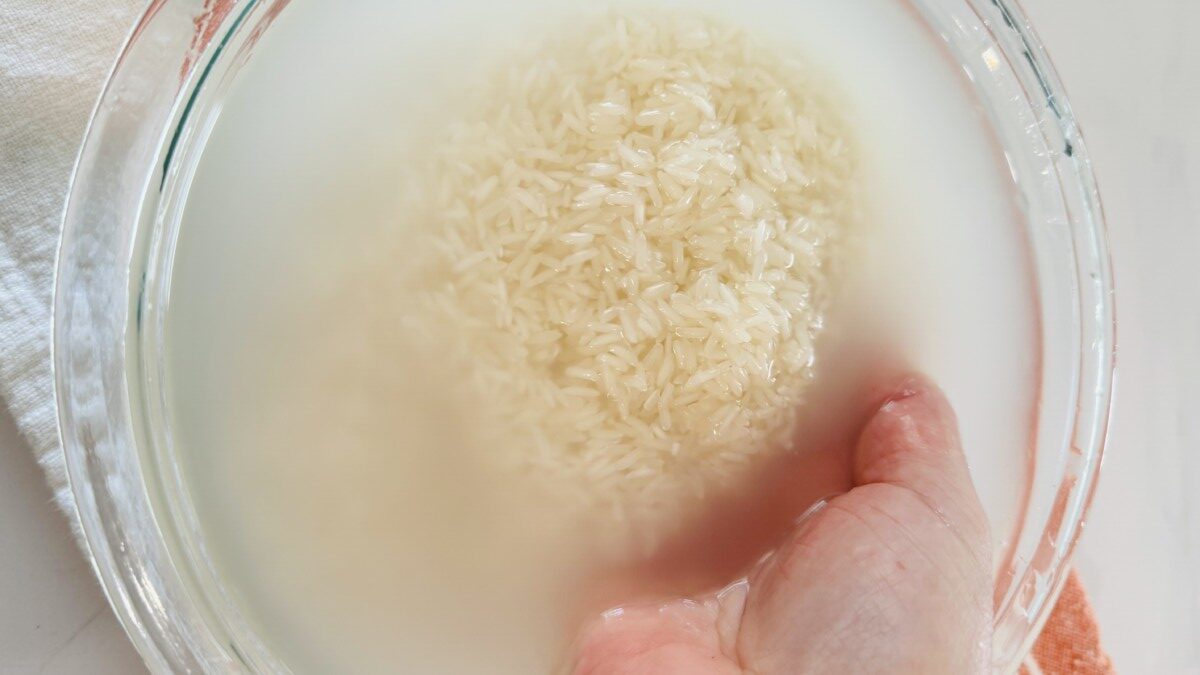 White basmati rice in a strainer under a running faucet, with clear water flowing through. A bowl of water sits beside the strainer.