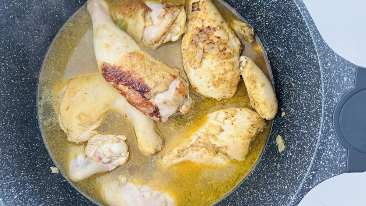 Chicken pieces and browned onions simmering in a pot filled with golden liquid. A sprig of fresh parsley or another herb rests on top.
