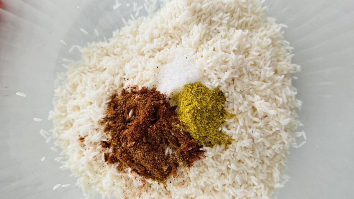 White basmati rice in a strainer positioned over a bowl. Spices like cardamom pods, cloves, and bay leaves are scattered on top of the rinsed rice.