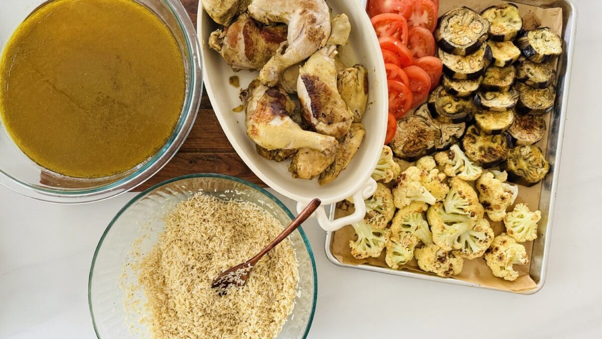 A colorful display of ingredients prepped and ready for layering in a Maqluba dish. Roasted eggplant slices, cauliflower florets, browned chicken pieces, a pot of simmering broth, and a bowl of seasoned rice with colorful spices visible are arranged on a table.