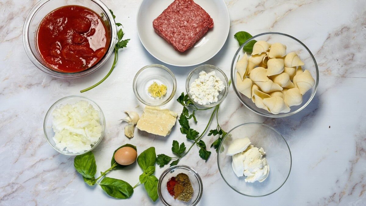 All ingredients for stuffed pasta shells measured and organized, including pasta shells, ground meat, cheeses, spices, and crushed tomatoes.