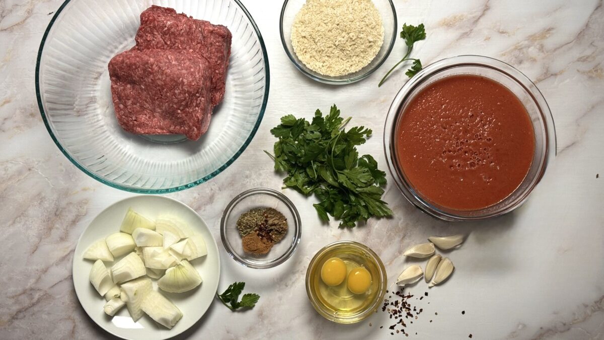 Measured ingredients for Greek meatballs, including ground beef, onions, garlic, panko breadcrumbs, parsley, eggs, and spices arranged on a countertop.