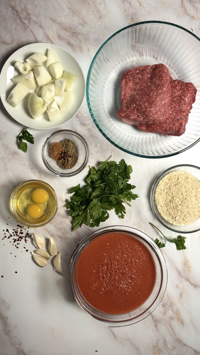 Measured ingredients for Greek meatballs, including ground beef, onions, garlic, panko breadcrumbs, parsley, eggs, and spices arranged on a countertop.