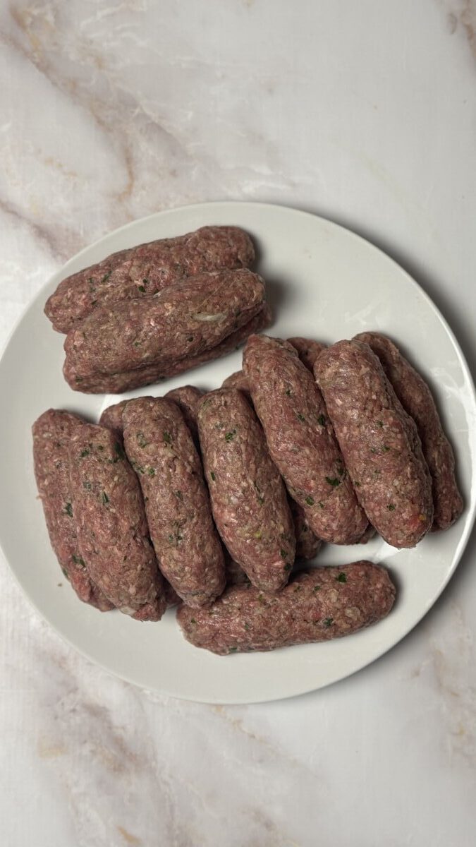 Hands shaping the Greek meatball mixture into sausage-like ovals on a clean surface.