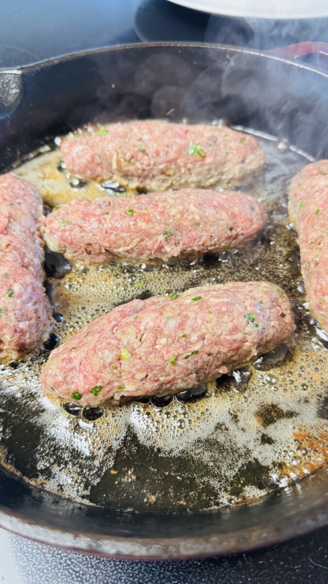 Greek meatballs being browned in a skillet with olive oil, developing a golden crust on each side.