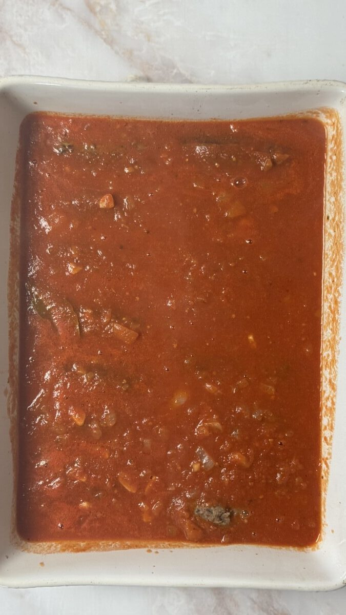 Rich tomato sauce being poured over Greek meatballs in a baking dish, ready to go into the oven.