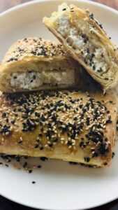 One whole piece of phyllo-wrapped baked feta on a plate with another piece cut in half, revealing the creamy feta filling and crispy phyllo layers.
