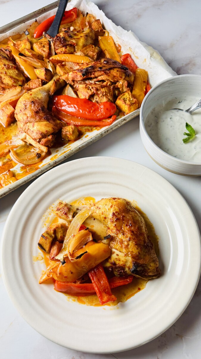 Harissa-roasted chicken and vegetables served on a plate with a side of creamy yogurt sauce, with the full baking sheet in the background.