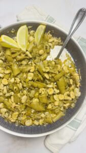 A pan of perfectly cooked fresh fava beans with a spoon, garnished with lemon slices and cilantro, ready to be enjoyed.