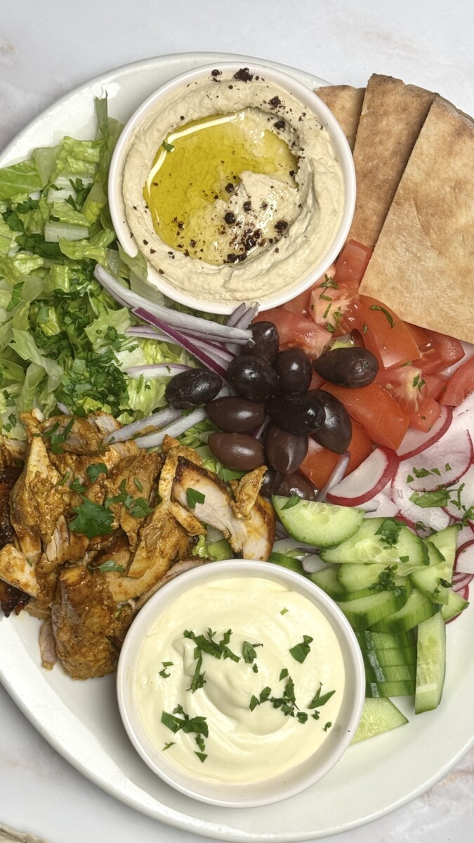 A vibrant chicken shawarma platter with hummus, tahini, tomatoes, olives, cucumber, and warm pita, served for a delicious Mediterranean feast.