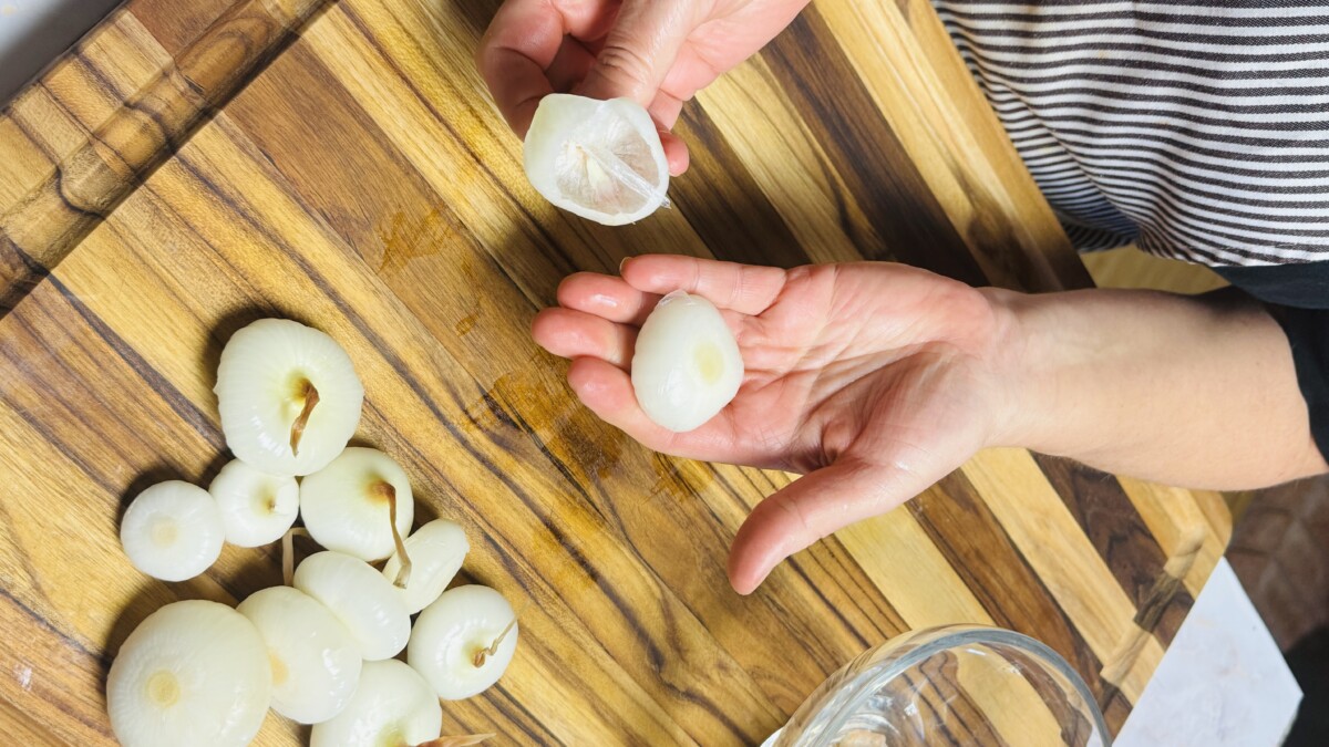 Hands peeling cipollini onions after simmering, removing their skins easily for Pomegranate Roasted Chicken.