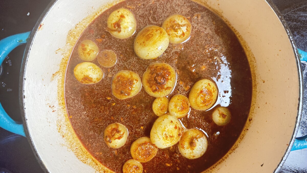 Pouring pomegranate broth into a pot with sautéed cipollini onions, creating a rich base for Pomegranate Roasted Chicken.