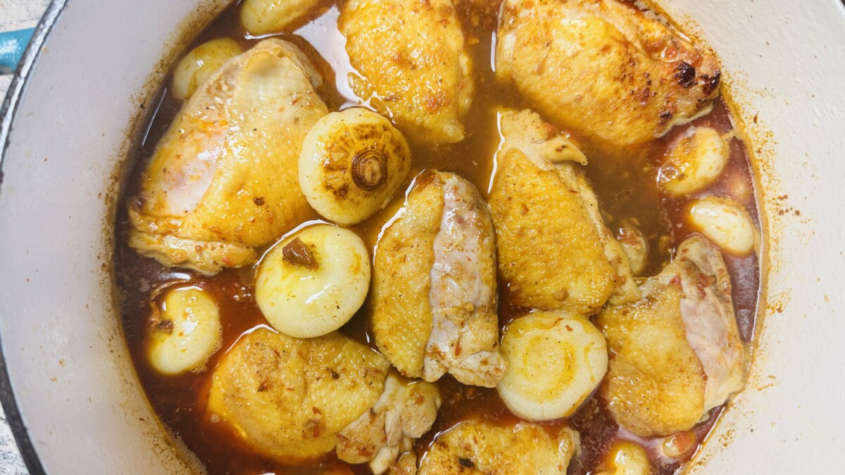 Pouring pomegranate broth into a pot with sautéed cipollini onions, creating a rich base for Pomegranate Roasted Chicken."