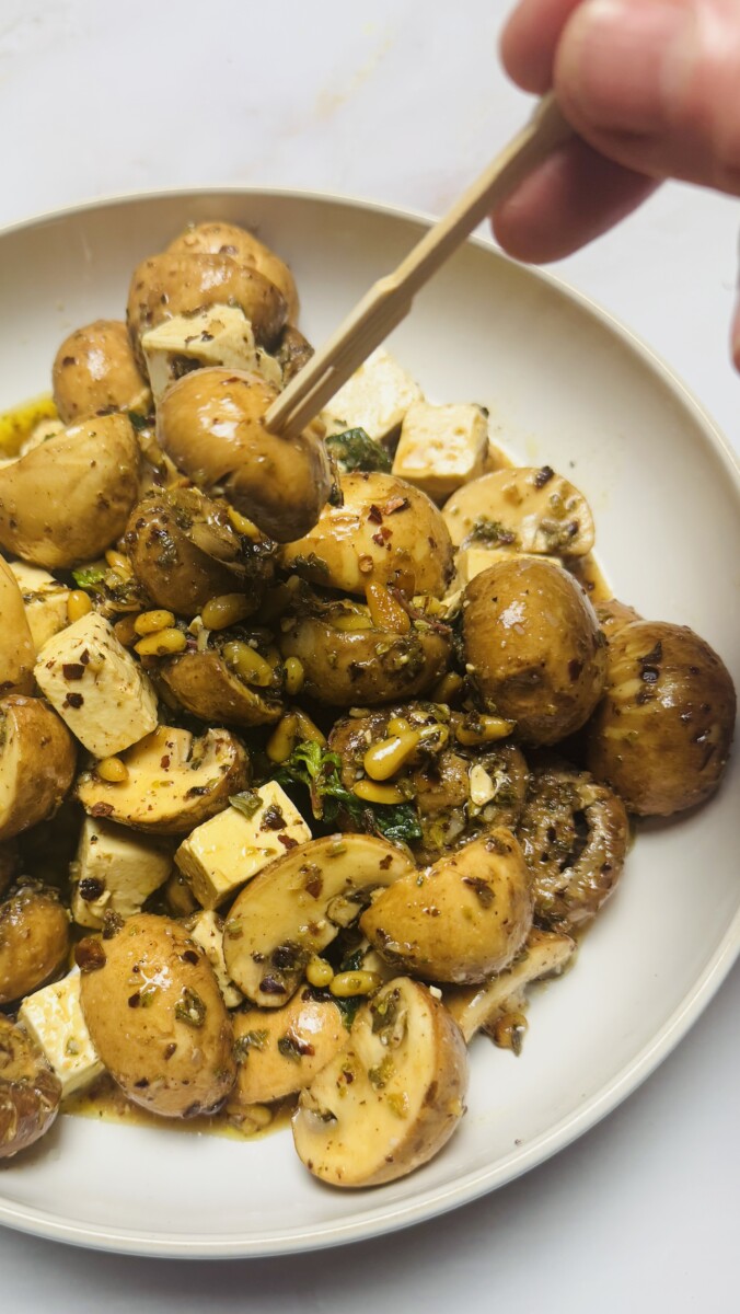 Mediterranean marinated mushrooms with za’atar and feta, served on a plate, with one mushroom held by a toothpick ready to enjoy.
