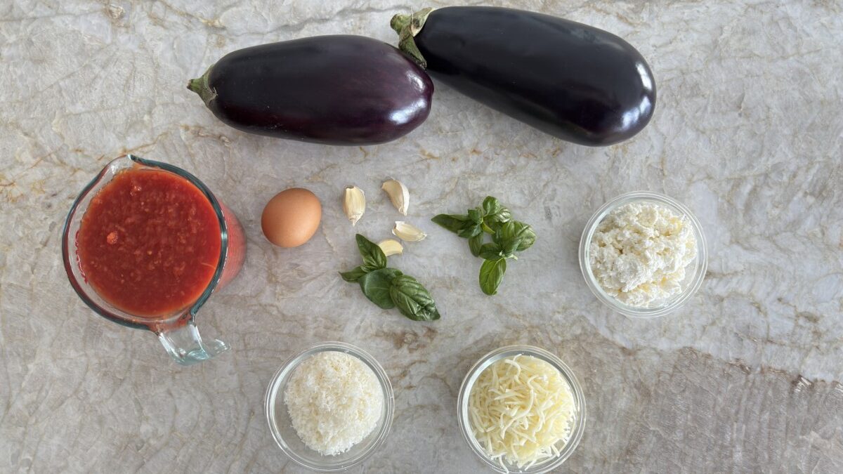 Ingredients for Eggplant Rollatini including eggplant, ricotta, marinara sauce, garlic, basil, mozzarella, parmesan, and egg, arranged on a kitchen counter.