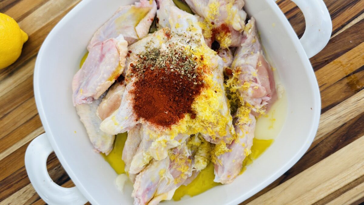 Chicken wings in a bowl topped with lemon zest, garlic, and spices, ready to be mixed for marination.