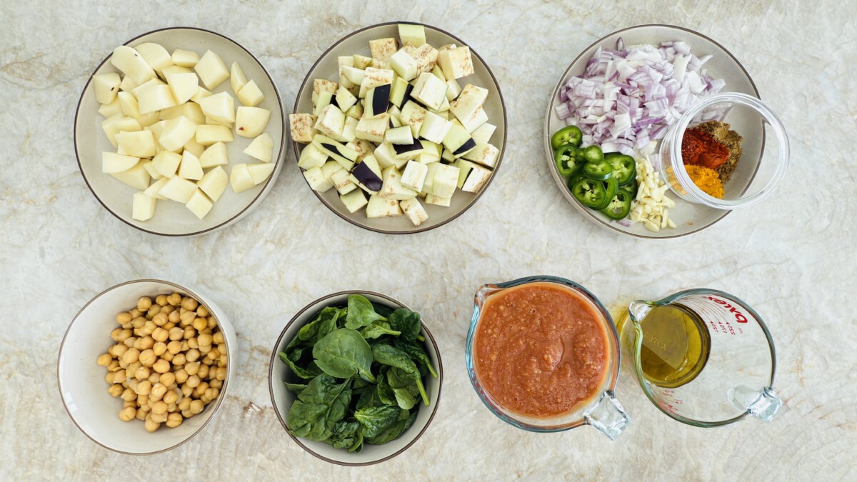 Fresh ingredients for Mediterranean eggplant stew, including cubed eggplant, potatoes, chickpeas, spinach, onion, garlic, tomato sauce, and spices.
