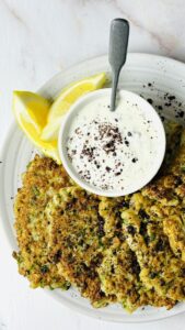 Golden cauliflower fritters served on a plate with a side of creamy tzatziki sauce and lemon wedges.