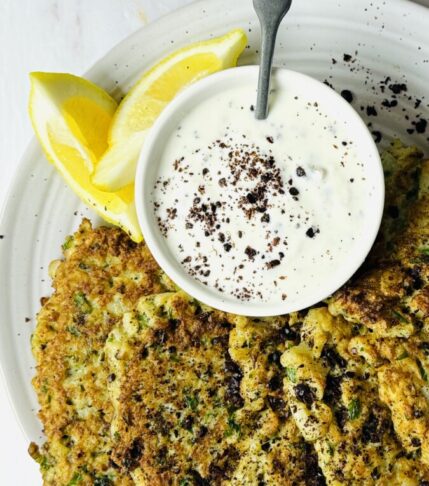 Golden cauliflower fritters served on a plate with a side of creamy tzatziki sauce and lemon wedges.