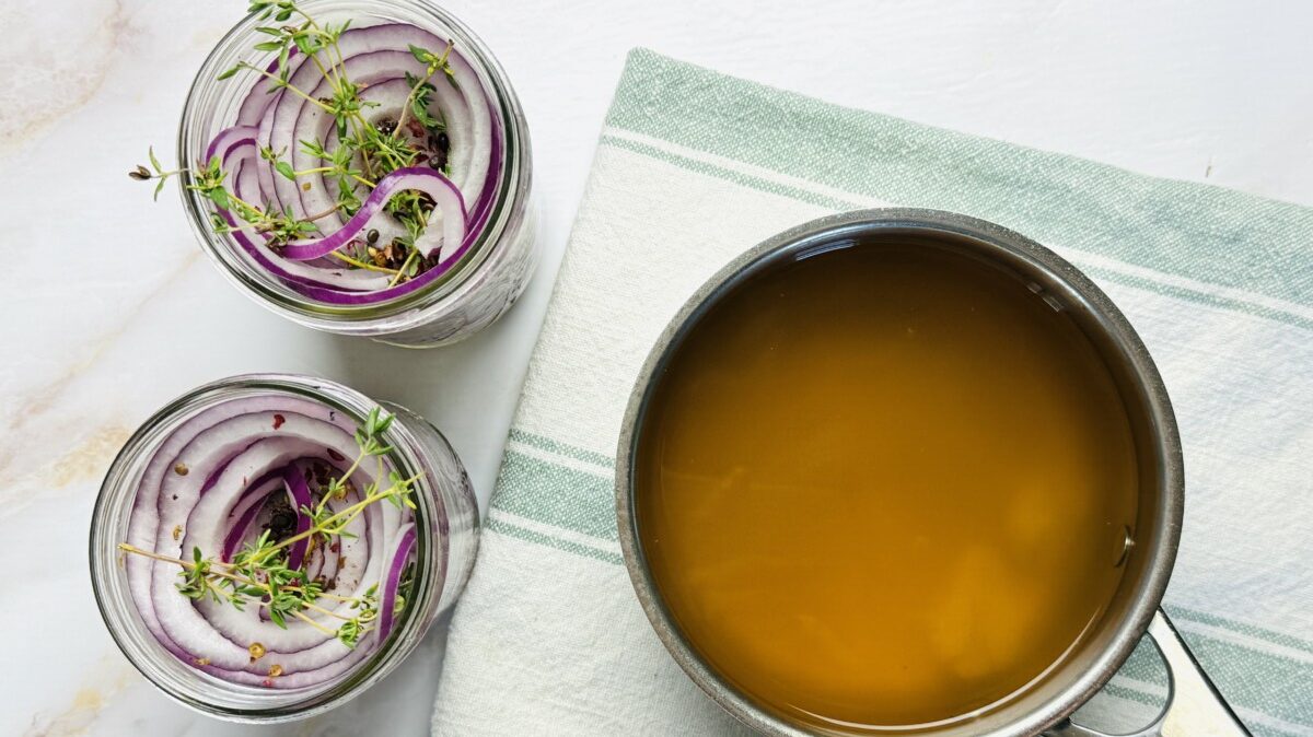Thinly sliced red onions packed in two jars with garlic cloves and thyme on top, next to a saucepan of hot brine.
