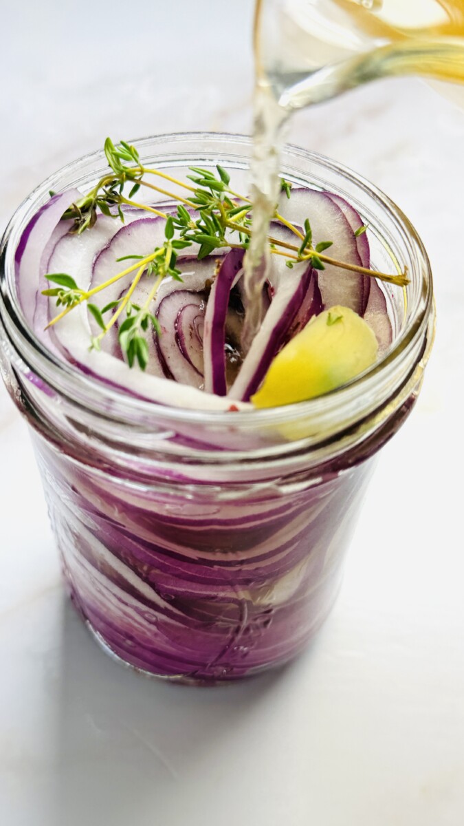 Hot pickling brine being poured from a saucepan into a jar filled with sliced red onions, garlic, and thyme.