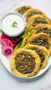A few baked sfeeha arranged on a serving platter with yogurt sauce and pickled vegetables on the side.