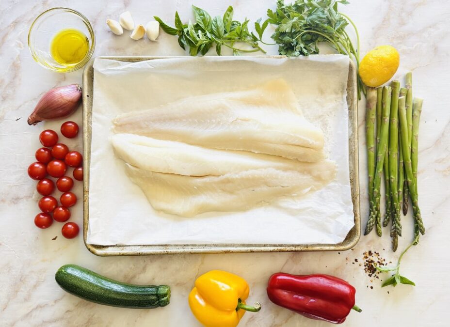 Ingredients for baked cod with vegetables, including cod fillets, zucchini, peppers, tomatoes, garlic, lemon, and fresh herbs
