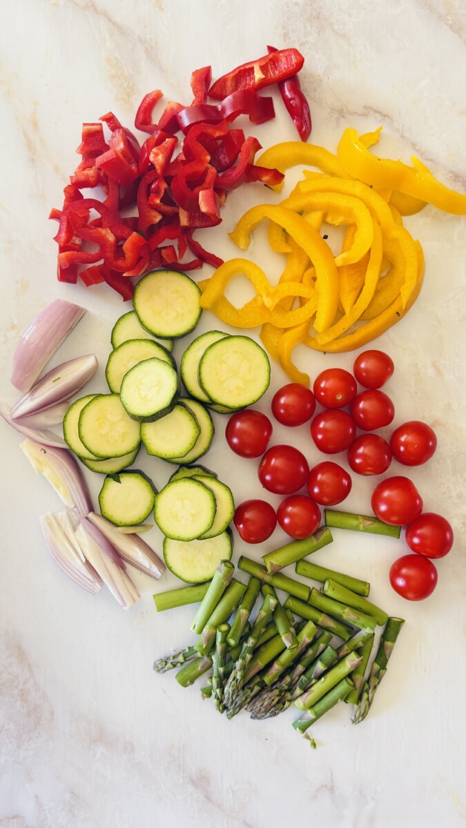 Sliced zucchini, bell peppers, cherry tomatoes, garlic, and shallots arranged on a cutting board