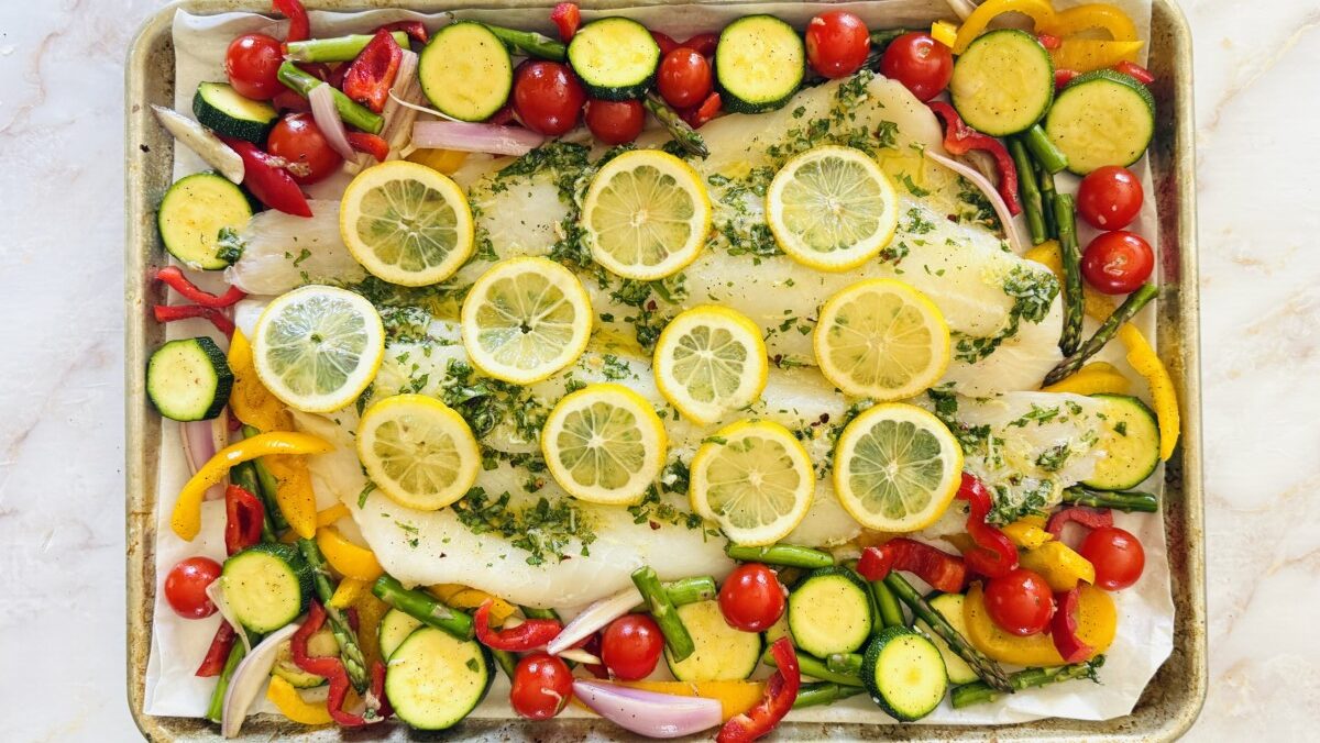 Cod fillets on a sheet pan surrounded by seasoned vegetables and topped with lemon slices, ready for the oven