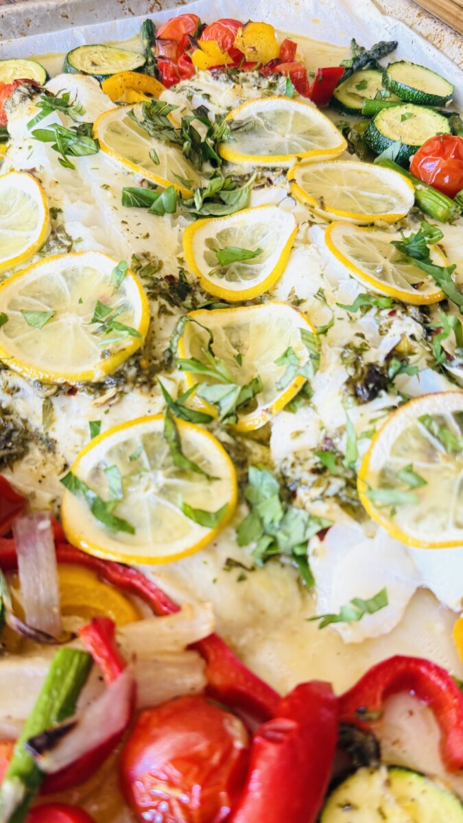 Baked cod fillets served with roasted vegetables and fresh herbs on a platter, garnished with lemon